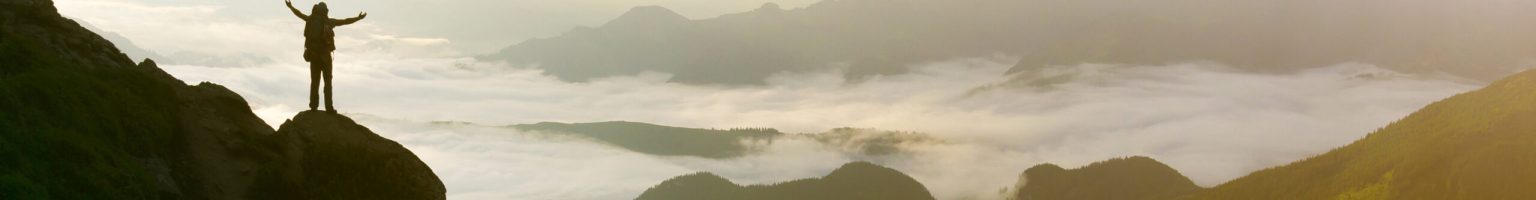 Wide mountain panorama. Small silhouette of tourist with backpack on rocky mountain slope with raised hands over valley covered with white puffy clouds. Beauty of nature, tourism and traveling concept